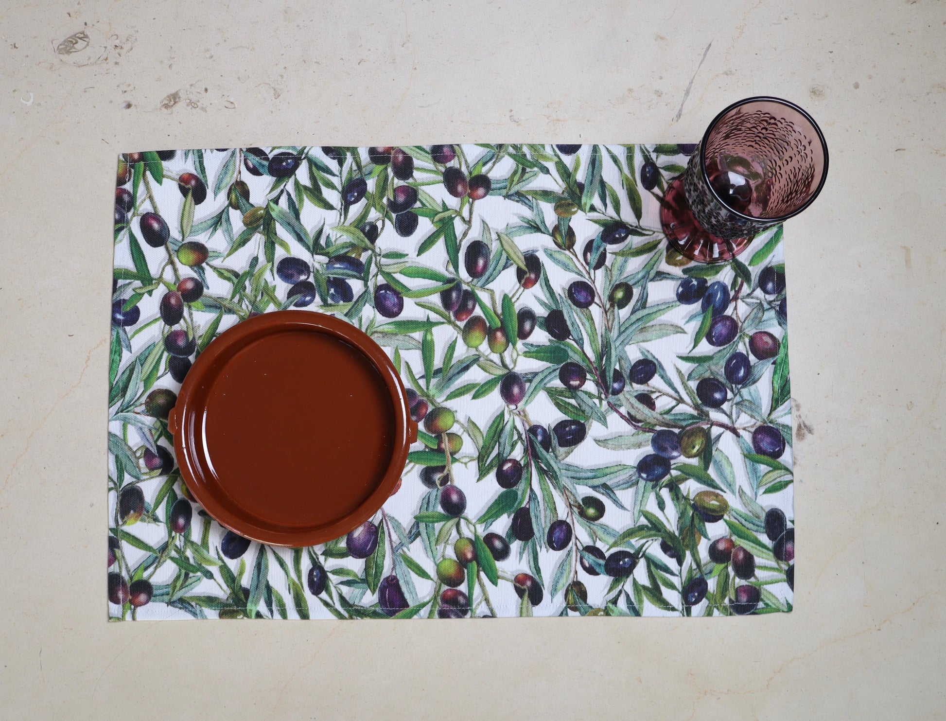 Mantel individual con estampado de aceitunas sobre mesa con plato terracota y copa.

